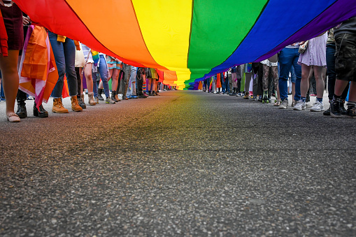 Under a LGBT Pride Flag with the legs of people carrying the flag with more copy space on road