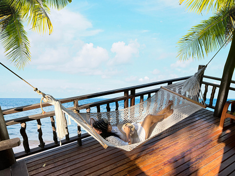 Happy woman tourist lying in hammock swing on wooden terrace over summer beach background. Happy holiday and relaxation concept.