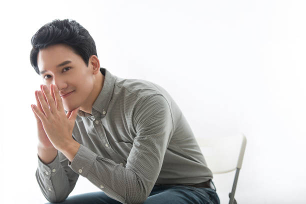 Young East Asian business man leaning forward in his chair, leaning on his elbow on his leg and resting his chin on his palm - stock photo stock photo