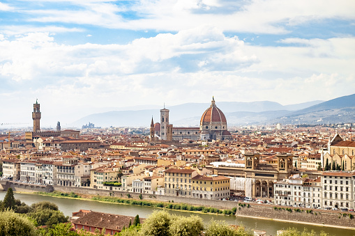 A skyline view of the city of Florence in Italy on a summer’s day, featuring the Cathedral of Santa Maria del Fiore. The Arno River runs through the foreground and a mountain range is seen in the distance.