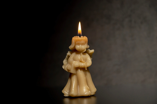 Wax candle in the shape of an angel, burning on a gray background.