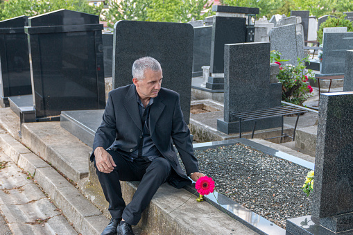 Mature man in black clothes on cemetery, holding a flower and Mourning for family loss. Concept for Death, Mourning, Funeral and Spirituality.