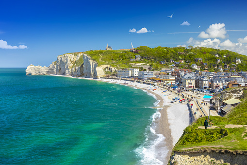Panoramic view of the beach of Etretat in Normandy, a popular french seaside town known for its chalk cliffs, with Notre-Dame de la Garde chapel overlooking the Amont cliff on a sunny day