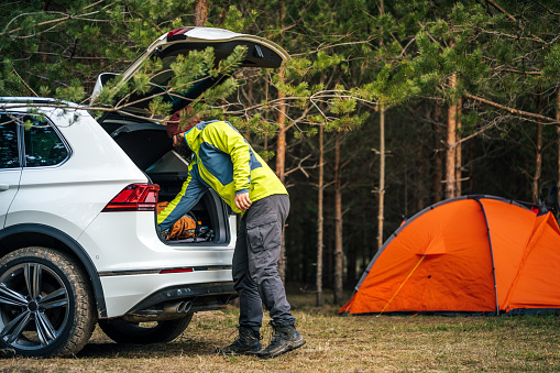 Weekend activities. Adult man arrived to the camp and getting equipment from the car trunk. An orange tent among the pine trees on the background.