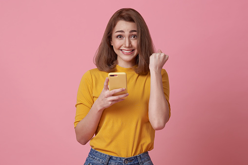 Happy young woman using mobile phone and hand gesture success celebrate isolated over pink background.