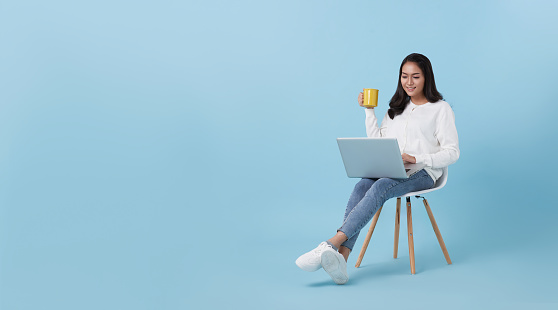 Young woman asian happy smiling in casual white cardigan with denim jeans.While her using computer laptop sitting on white chair and drinking coffee isolate on bright blue background.