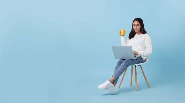 Young woman asian happy smiling in casual white cardigan with denim jeans.While her using computer laptop sitting on white chair and drinking coffee isolate on bright blue background. stock photo