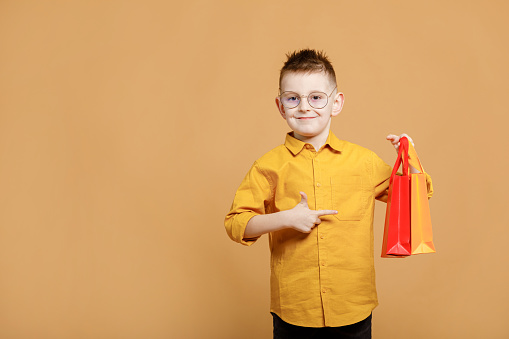 Shopping on black friday. Little boy holding shopping bags on yellow background and showing finger on bag. Shopper with many colored paper bags. Holidays sales and discounts. Cyber Monday