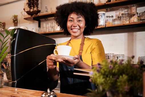African American female barista in looks at camera, offers cup of coffee to customer with cheerful smile, happy service works in casual restaurant cafe, young small business startup entrepreneur.