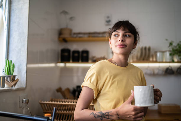 joven contemplativa bebiendo café o té y mirando la ventana de casa - cándido fotografías e imágenes de stock