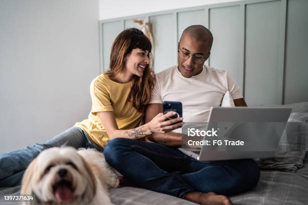 Young woman showing something to husband on the mobile phone with the dog in the bed at home