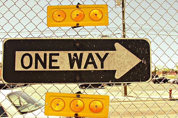 A one way sign hanging on a chain link fence in a paved-over suburban area.