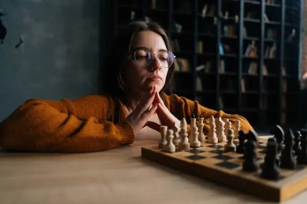 Thoughtful cute young woman in elegant eyeglasses thinking about chess move sitting on floor in dark library room, selective focus. Pretty intelligent lady playing logical board game alone at home.
