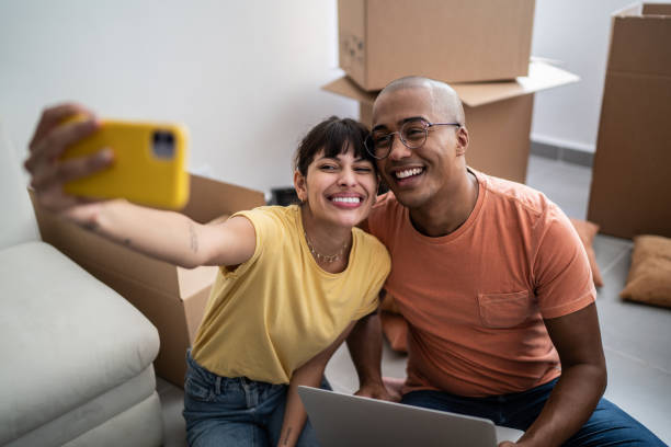 Young couple taking a selfie or filming using mobile phone while packing moving boxes at home