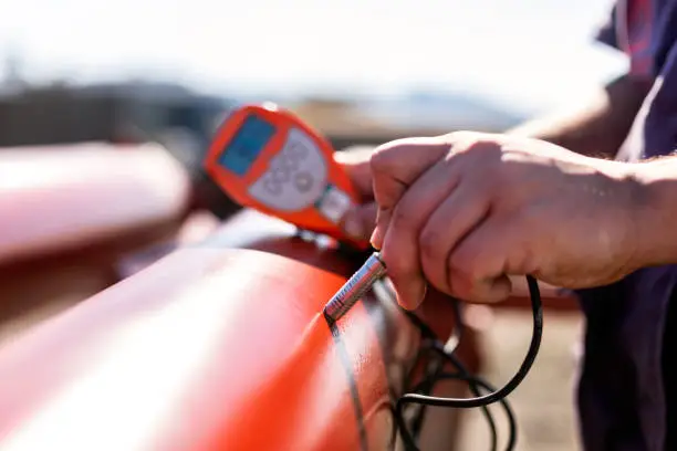 Paint inspector is measuring to red colored paint coating of the pipe in construction site. A coating is a covering that is applied to the surface of an object, usually referred to as the substrate.