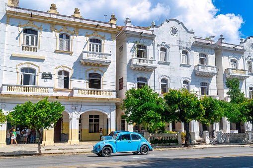 Havana, Cuba - May 24, 2022: Incidental people are out and about in an avenue in the Vedado district. A VW beetle vintage car drives by the old buildings in the area