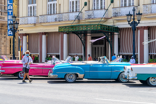 Havana, Cuba - May 24, 2022: A group of people are in their daily routines by the Hotel Inglaterra in the Parque Central district. Vintage cars working as tourist taxis are parked in front of the resort.