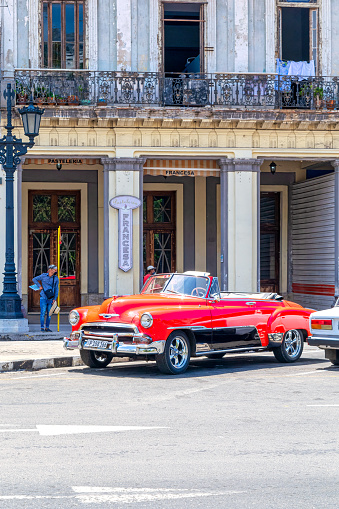 Havana, Cuba - May 24, 2022: A man leans on a traffic signal by a building reading 'Francesa'. A vintage convertible car is parked in the street. The upper part of the building is run-down and weathered