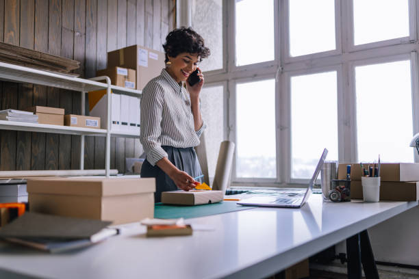 una mujer de negocios feliz hablando en su teléfono inteligente mientras prepara paquetes para el envío en su tienda - entrepreneur fotografías e imágenes de stock