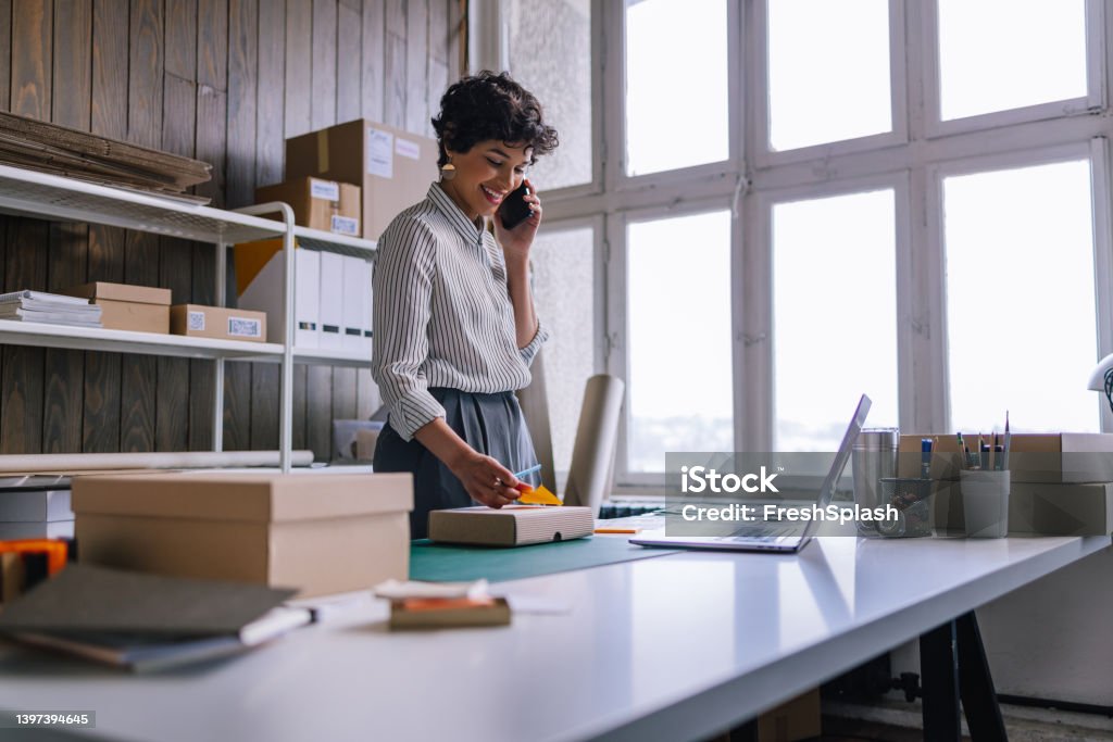 A Happy Businesswoman Talking On Her Smartphone While Preparing Packages For Shipping  In Her Store A beautiful smiling female Latin-American entrepreneur using her mobile phone while packing merchandise for delivery  in cardboard boxes. Entrepreneur Stock Photo