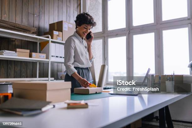 A Happy Businesswoman Talking On Her Smartphone While Preparing Packages For Shipping In Her Store Stock Photo - Download Image Now