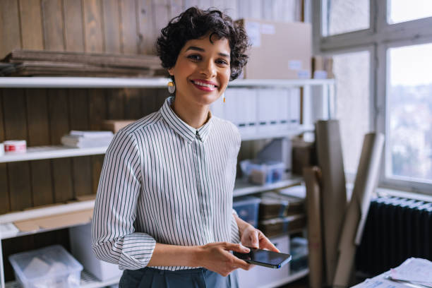 uma empresária sorridente olhando para a câmera enquanto agendava um compromisso de entrega em seu smartphone - warehouse manager place of work portrait - fotografias e filmes do acervo