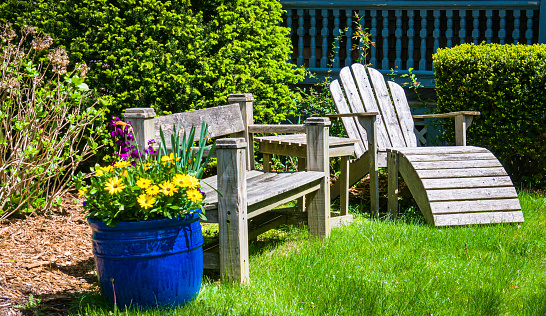 A wooden bench and an  Adirondack chair rest next to a blue ceramic pot full of springtime flowers in a Cape Cod garden.