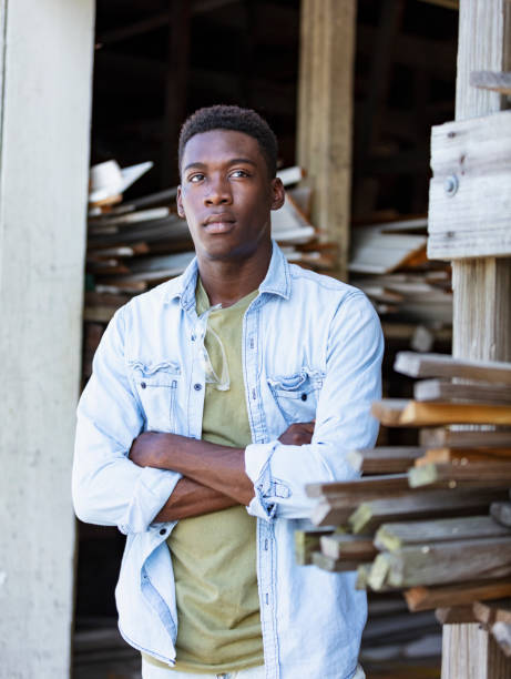 A young African-American man working at a lumberyard, standing by a storage area. He has a serious expression on his face, arms crossed, looking slightly away from the camera.