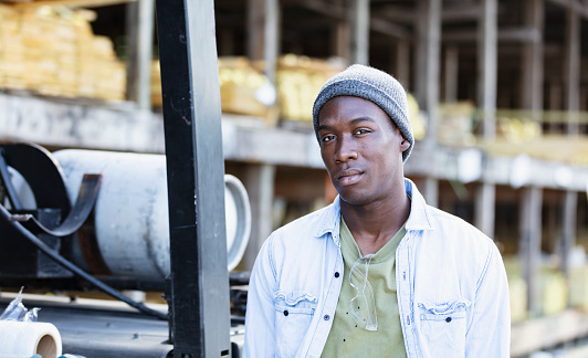 A young African-American man working in a lumberyard or building supply store, looking toward the camera, standing next to a forklift. Shelves full of lumber are out of focus in the background.