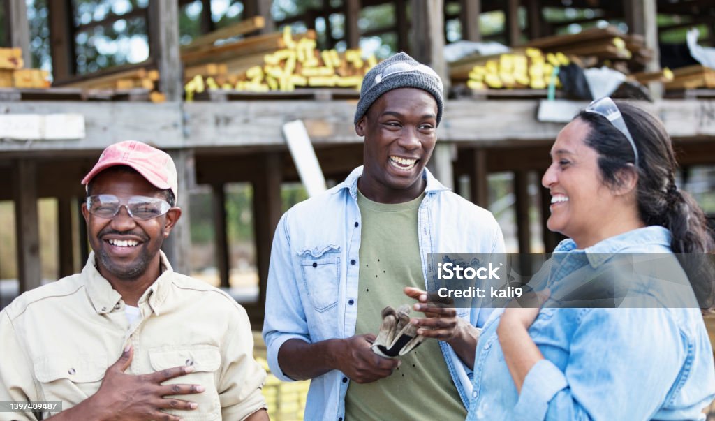 Multiracial group of workers at lumberyard, laughing A multiracial group of three workers at a lumberyard, standing together, laughing. The Hispanic woman and the African-American man wearing the trucker's hat and safety glasses are in their 40s. The main focus is on the young African-American man standing in the middle. He is in his 20s. There are shelves stacked with lumber out of focus behind them. Blue-collar Worker Stock Photo