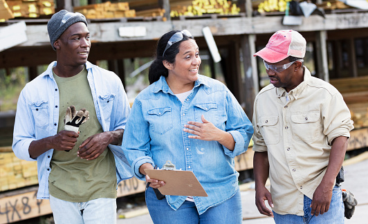 A multiracial group of three people working in a lumberyard. The manager is a mature Hispanic woman who is holding a clipboard. She is in the middle, looking toward the older African-American man, talking and smiling as he looks down at the clipboard.