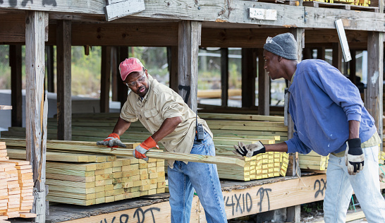 Two African-American men working together to put wood planks on outdoor shelves at a lumberyard or building supply store. Or, perhaps they are customers, buying lumber for a project. The focus is on the older man, in his 40s, wearing a trucker's hat. His assistant is a young man in his 20s.