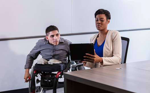 Two business people having a brainstorming session in an office meeting room. The young man is in a wheelchair, an amputee with spina bifida. His coworker is a mid adult African-American woman, sitting at the table next to him, holding up a laptop computer so they can both look at the screen.
