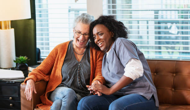 Senior woman at home with healthcare worker A senior woman in her 80s sitting on a sofa at home with a health care aide. The nurse is an African-American woman in her 30s wearing scrubs. She is holding the patient's hand, giving her physical and emotional support. They are both smiling. cheek to cheek photos stock pictures, royalty-free photos & images