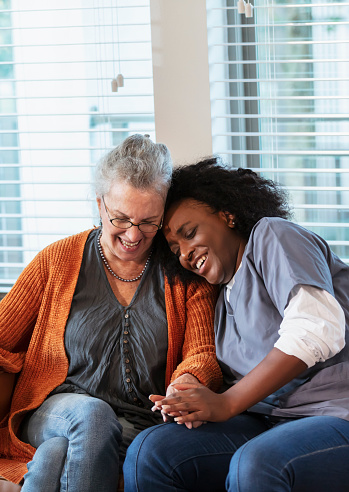A senior woman in her 80s sitting on a sofa at home with a health care aide. The nurse is an African-American woman in her 30s wearing scrubs. She is holding the patient's hand, giving her physical and emotional support. They are both smiling.