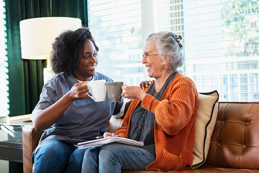 A senior woman in her 80s sitting on the living room couch, having coffee and conversing with a home healthcare aide, an African-American woman in her 30s.