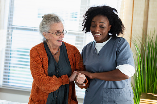 A home healthcare aide, an African-American woman in her 30s, helping a senior woman in her 80s walk. They are in the patient's home, standing by a sunny window. The healthcare worker is holding the patient's arm and hand for support as she tries to walk.