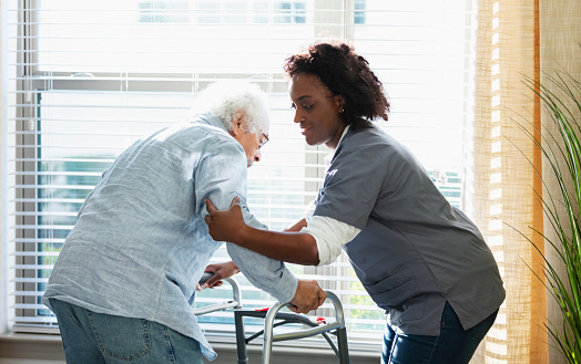 A home healthcare aide, an African-American woman in her 30s, helping a senior man in his 80s stand up from sitting on his bed and use a walker. He is Hispanic and Caucasian.