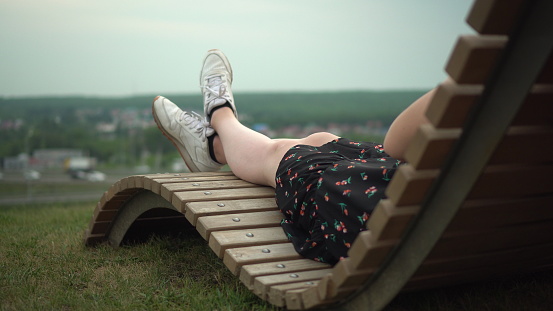 A young girl in a dress and sneakers lies on a deck chair on the grass. Girl's legs close up. 4k