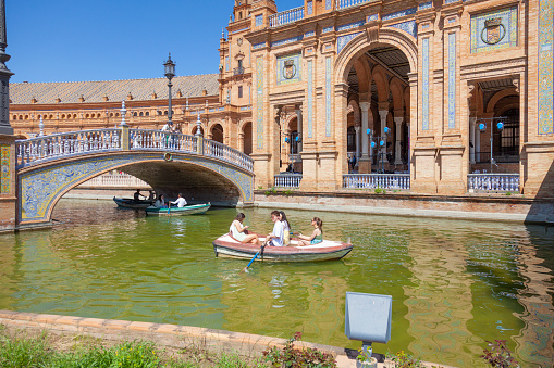 Seville, Spain - 7th April 2022 Visitors to the Plaza de España  in Seville, Andalucía on a sunny April afternoon. Some are rowing on the canal. A man and a woman are watching from a bridge. The Plaza de España was designed by Anibal Gonzalez and completed in 1929  as the Pabellon de Andalucia for Expo 29.