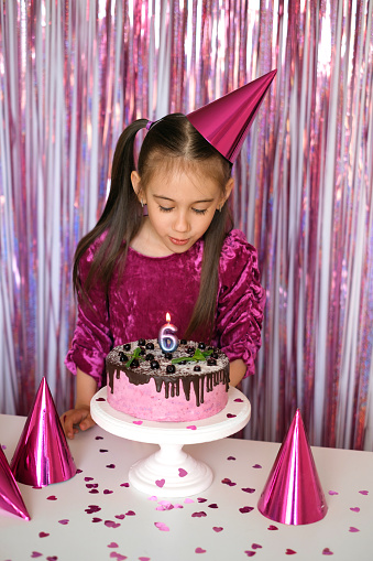 A cute little girl in a pink dress with a festive hat on her head blows out a candle in the shape of the number 6 on the cake. The child celebrates his birthday.