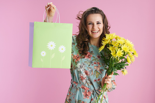 happy elegant woman with long wavy brunette hair with yellow chrysanthemums flowers and green shopping bag isolated on pink background.