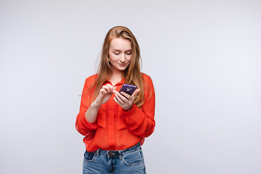 Front view of happy woman keeping phone and messaging on grey isolated background. Girl looking at smartphone, reading and texting. Concept of internet and modern technology.