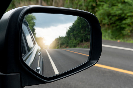Inside view of mirrors wing. Rear view of a gray car with asphalt road and green trees in the daytime. Clear traffic in rural areas.