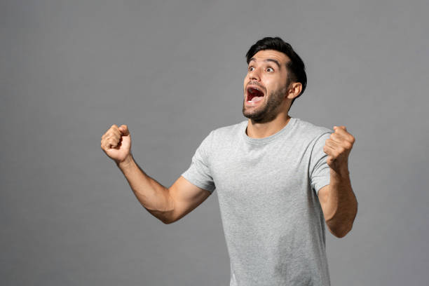 Gleeful Caucasian man with clenched fists Delighted Caucasian man in casual wear celebrating success with clenched fists while standing on gray isolated studio background exhilaration stock pictures, royalty-free photos & images