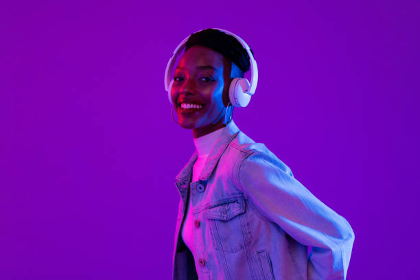 femme afro-américaine élégante et souriante portant des écouteurs et écoutant de la musique dans un fond de studio violet moderne - casque musique photos et images de collection