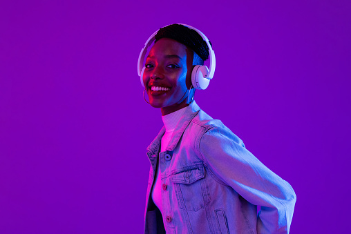 Stylish happy smiling African-American woman wearing headphones and listening to music in modern purple studio background