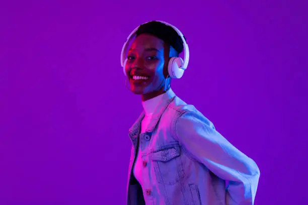 Photo of Stylish happy smiling African-American woman wearing headphones and listening to music in modern purple studio background