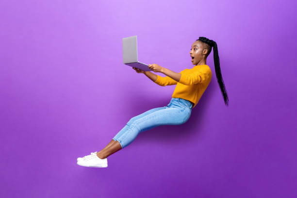 Astonished black woman levitating while working on laptop Full body side view of amazed young African American woman levitating while browsing netbook computer on purple background in light studio floating woman stock pictures, royalty-free photos & images