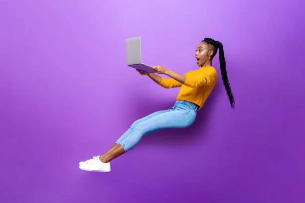 Full body side view of amazed young African American woman levitating while browsing netbook computer on purple background in light studio